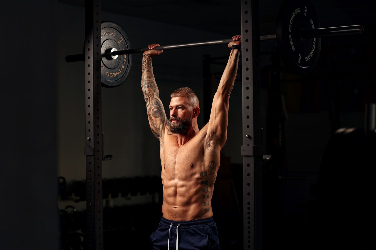 Athletic man lifting barbell overhead in dark gym, showing muscular physique and tattoos.