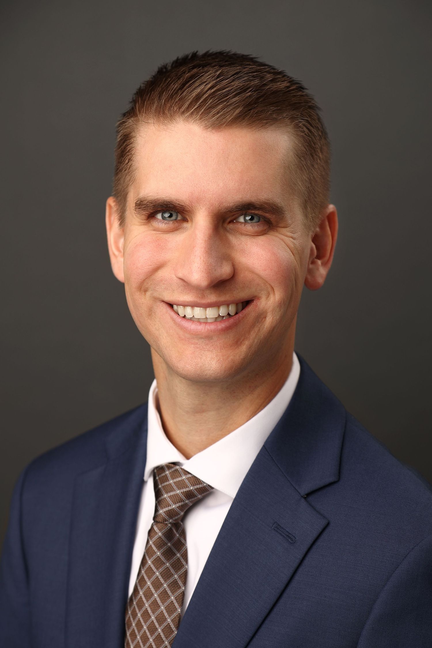 Professional headshot of smiling man in navy suit and tie against gray background.