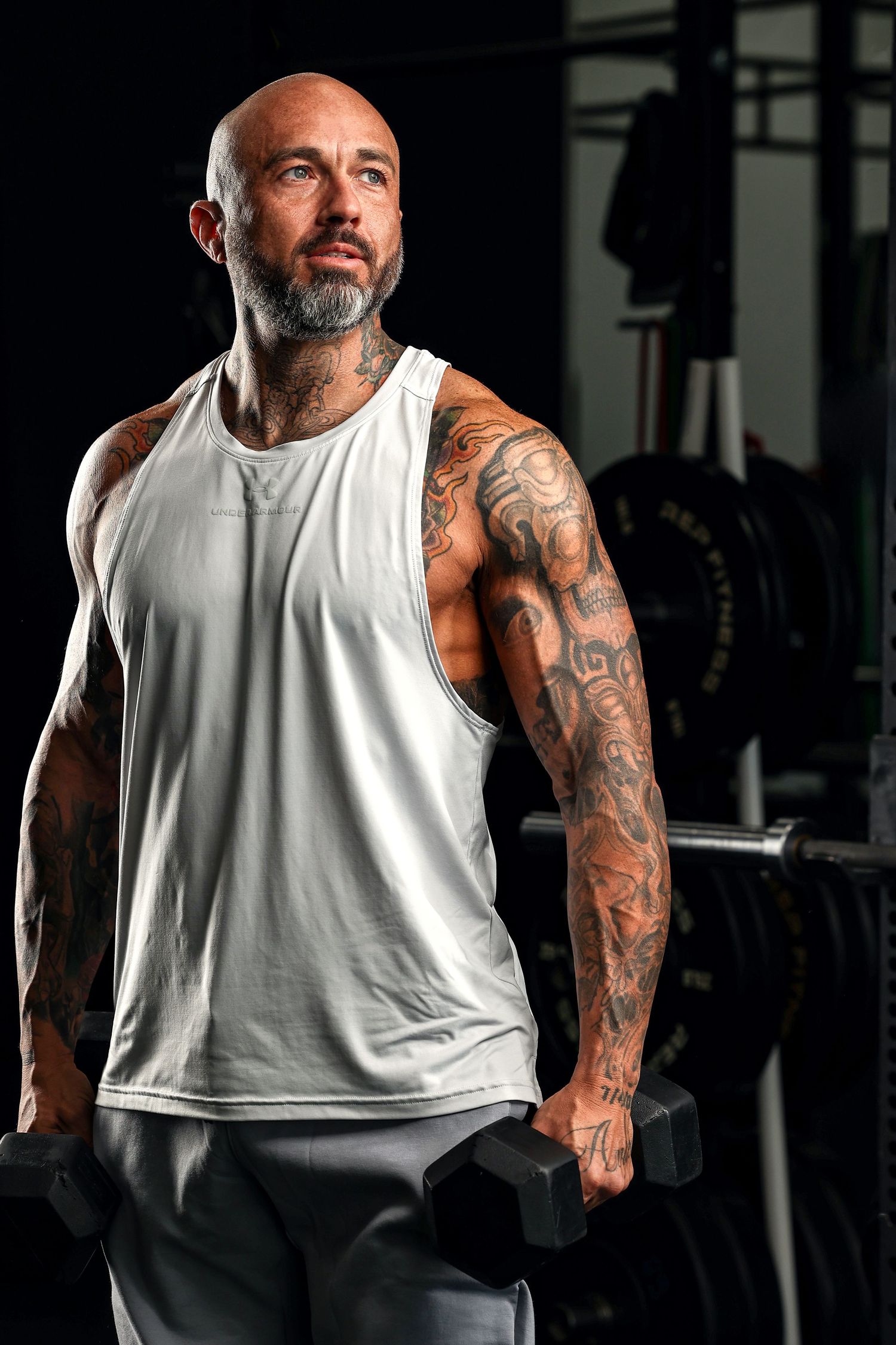 Tattooed bald man with beard lifting dumbbells wearing white tank top in gym.