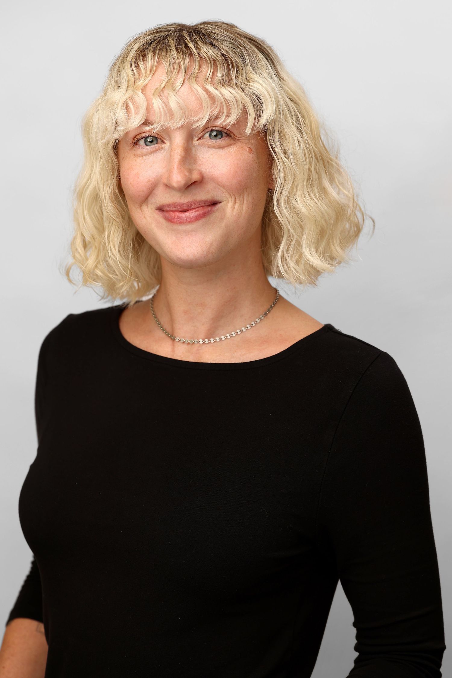 Professional headshot of blonde woman with curly bob wearing black top and silver necklace.