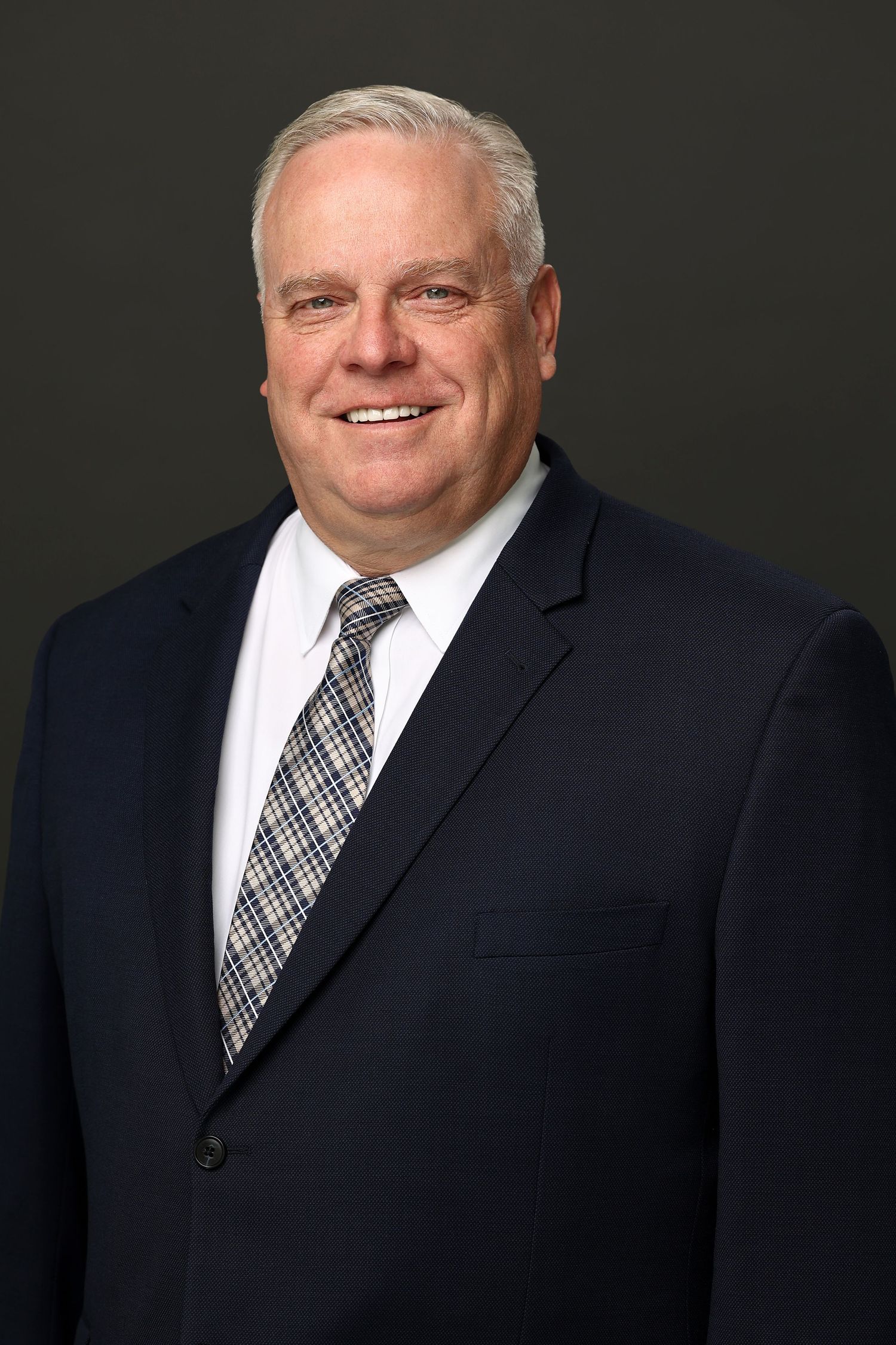 Professional headshot of a smiling older man in a navy suit and plaid tie against a dark background.