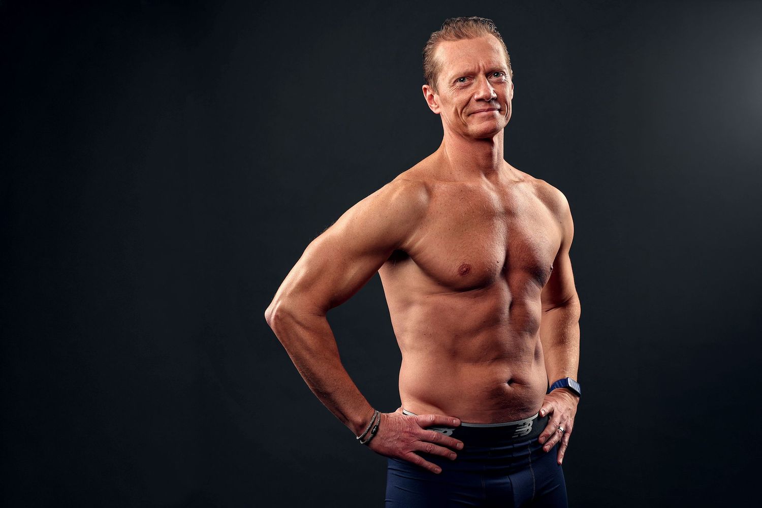 Shirtless muscular middle-aged man posing with hands on hips against dark background.