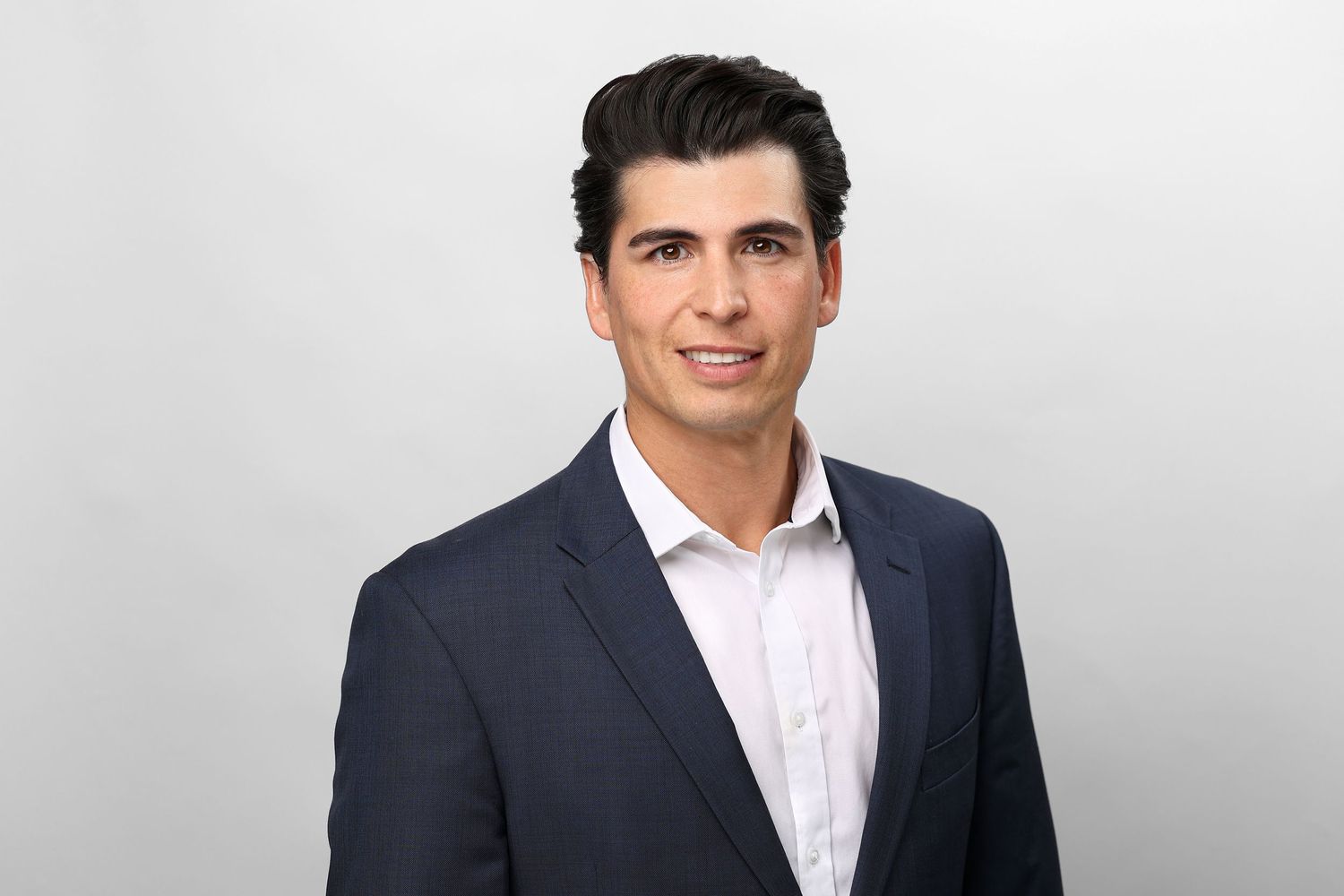 Professional headshot of a young man with dark hair in a navy blazer and white shirt on a light background.