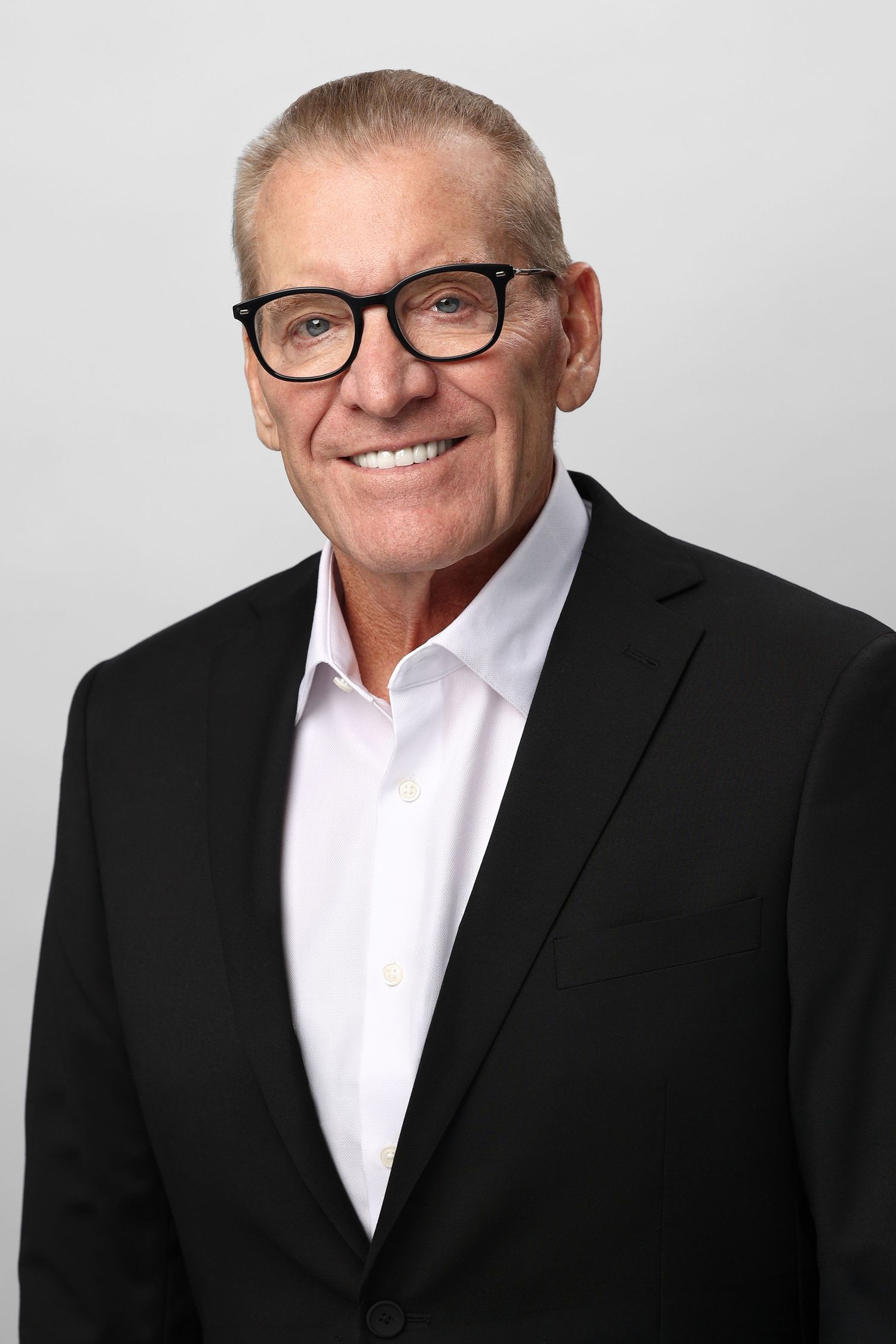 Senior businessman with glasses wearing black suit smiles for professional portrait.