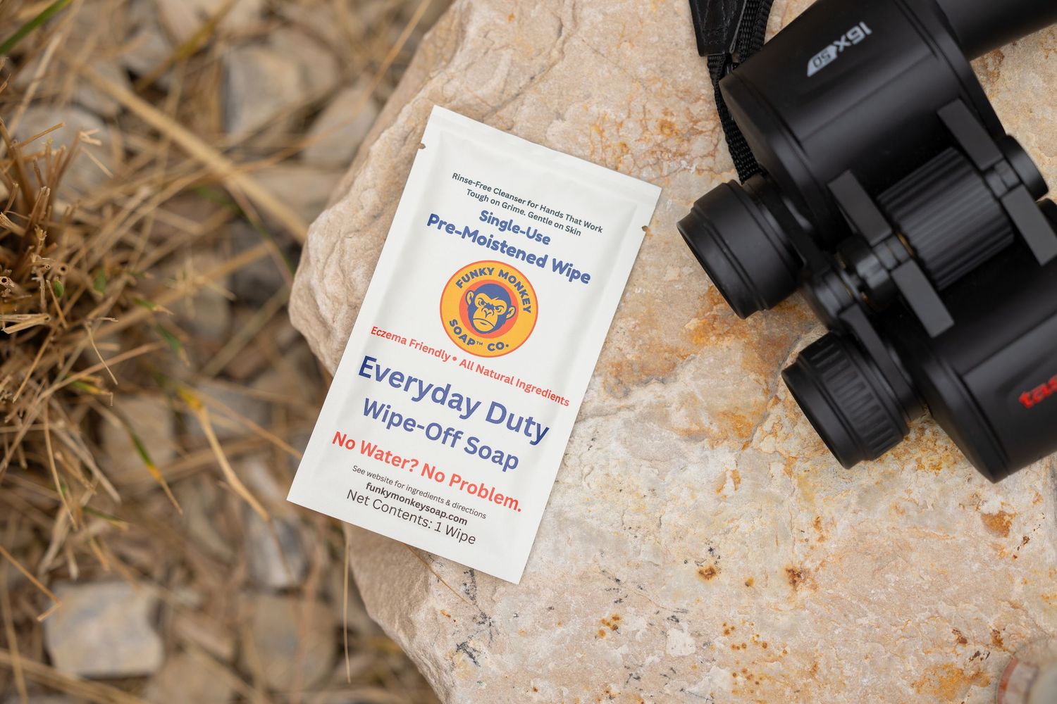 Everyday Duty Wipe-Off Soap packet resting on a rock next to binoculars outdoors.
