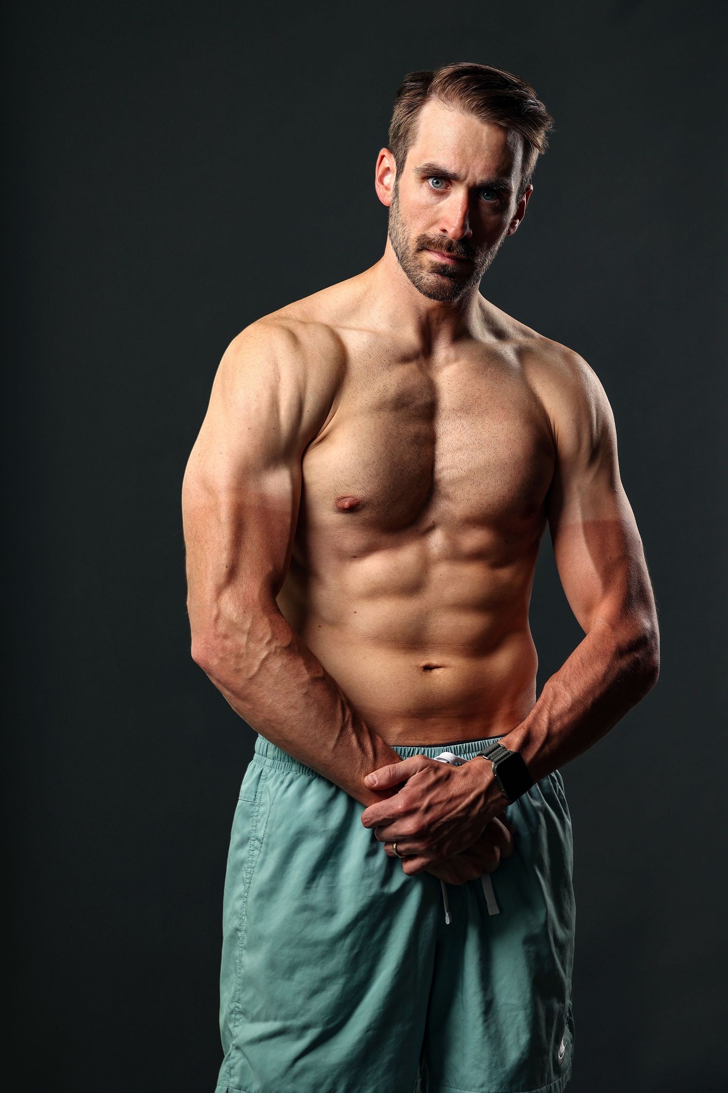 Fit shirtless man with six-pack abs standing confidently in teal shorts on dark background.