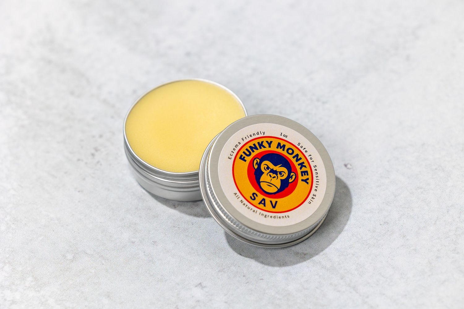 Open tin of Bunky Monkey SAV balm showing yellow salve on marble surface.