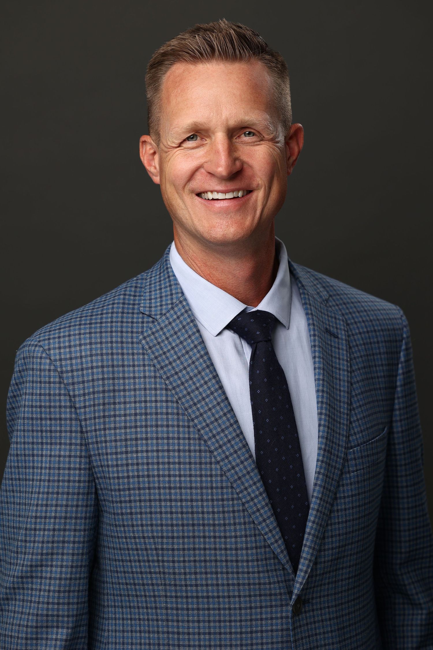 Professional headshot of tall blonde man in blue plaid blazer and tie smiling warmly.
