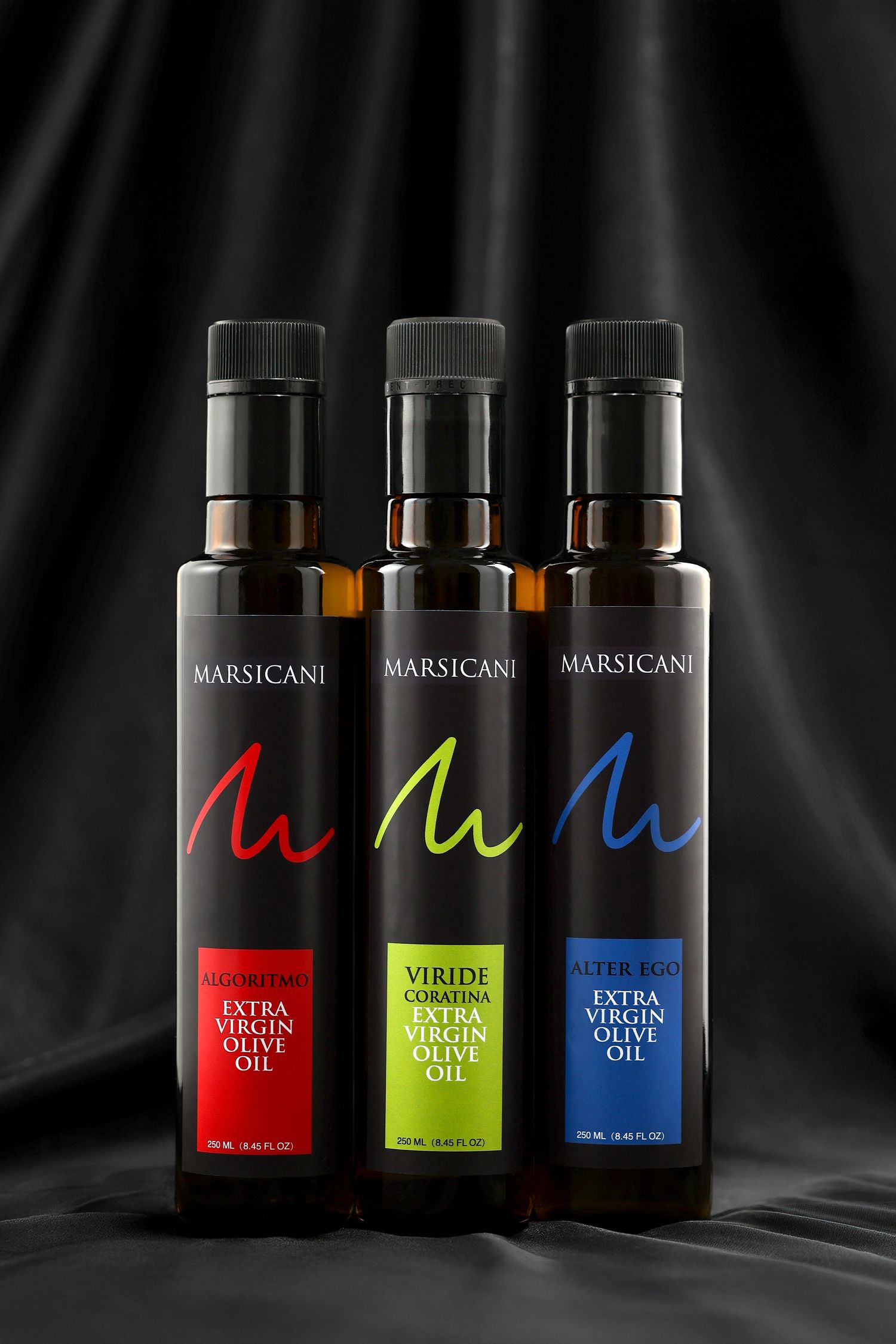 Three Marsicani extra virgin olive oil bottles with colorful labels on dark silk background.