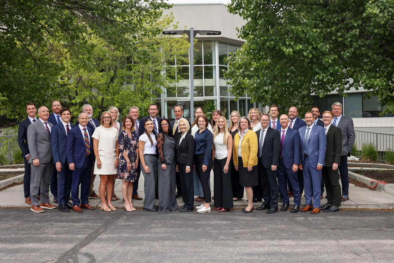 Large professional group photo of business team in formal attire posing outdoors near modern building.