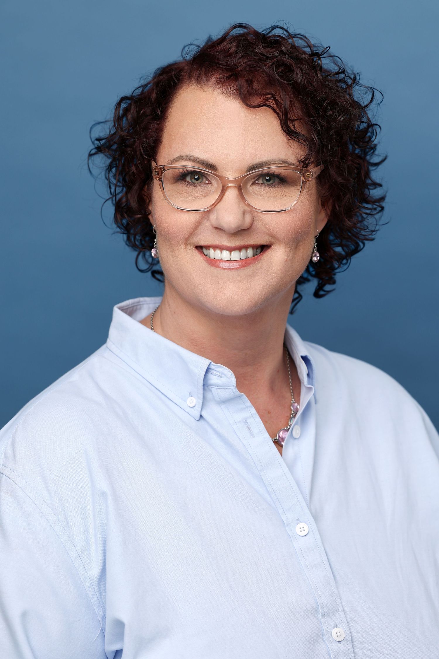 Professional headshot of a smiling woman with curly red hair and glasses wearing a light blue shirt.