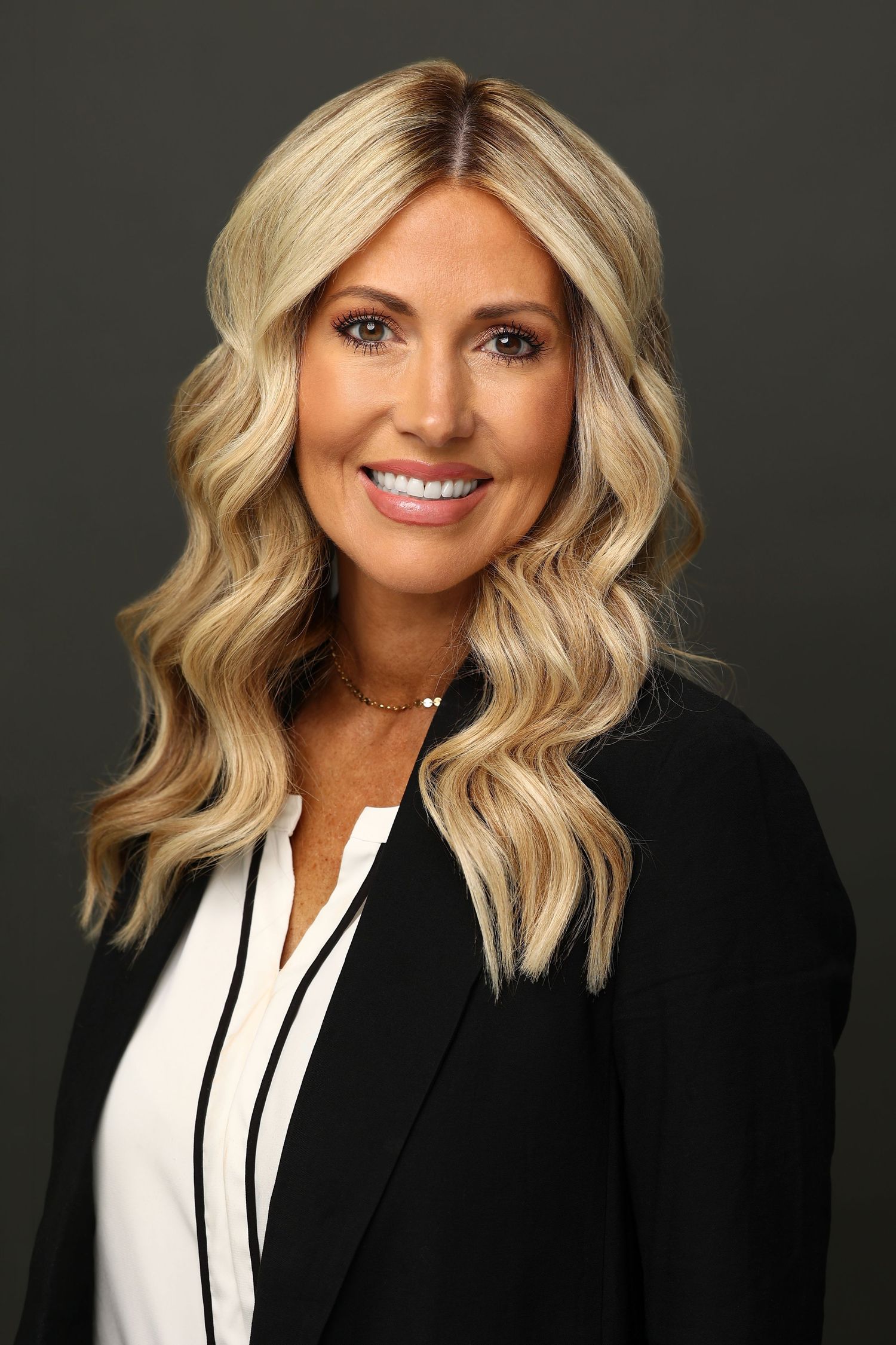 Professional headshot of blonde woman in black blazer and white blouse smiling confidently.