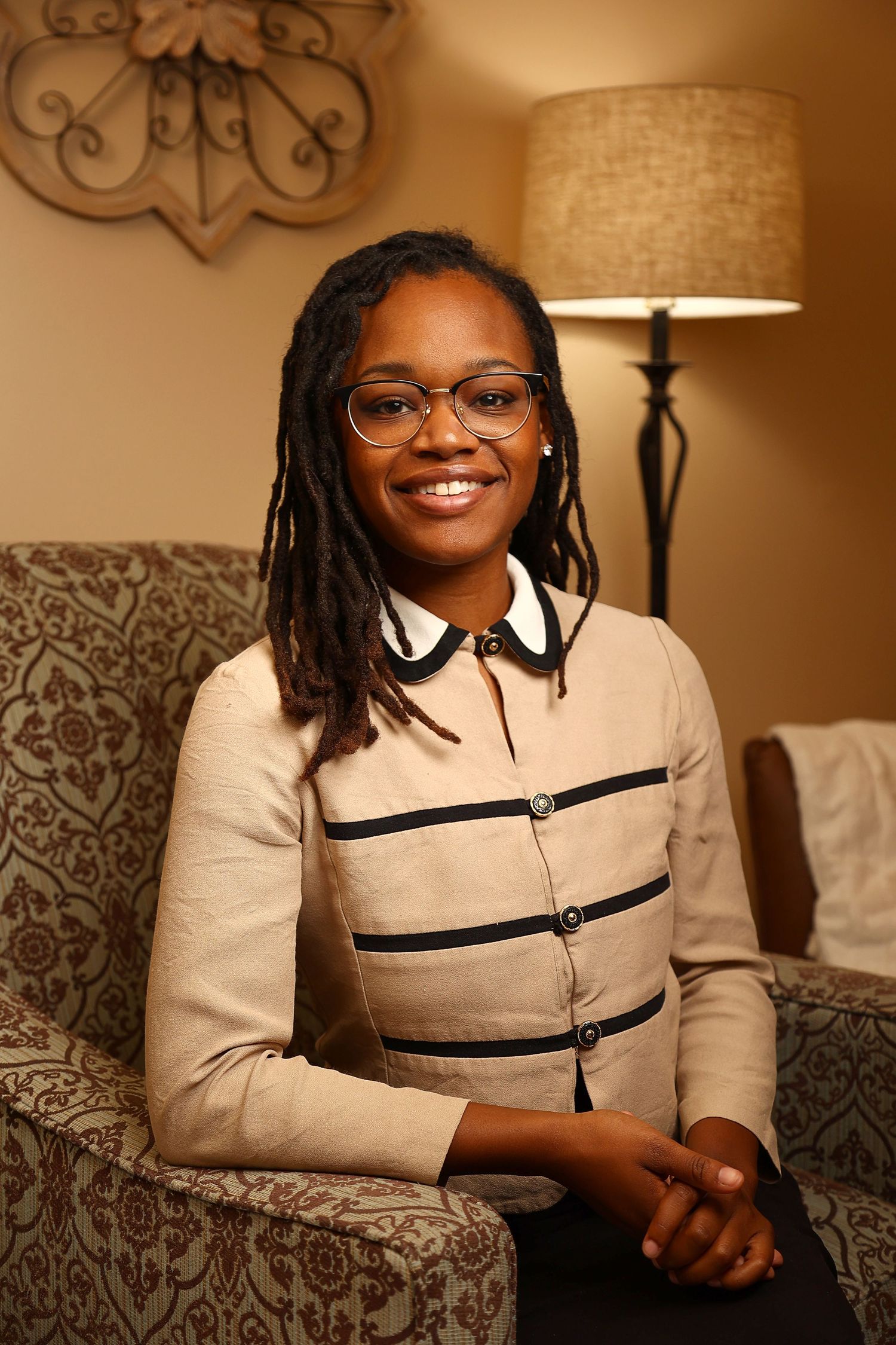 Smiling young woman with locs and glasses wearing a beige striped blazer seated in a cozy office setting.