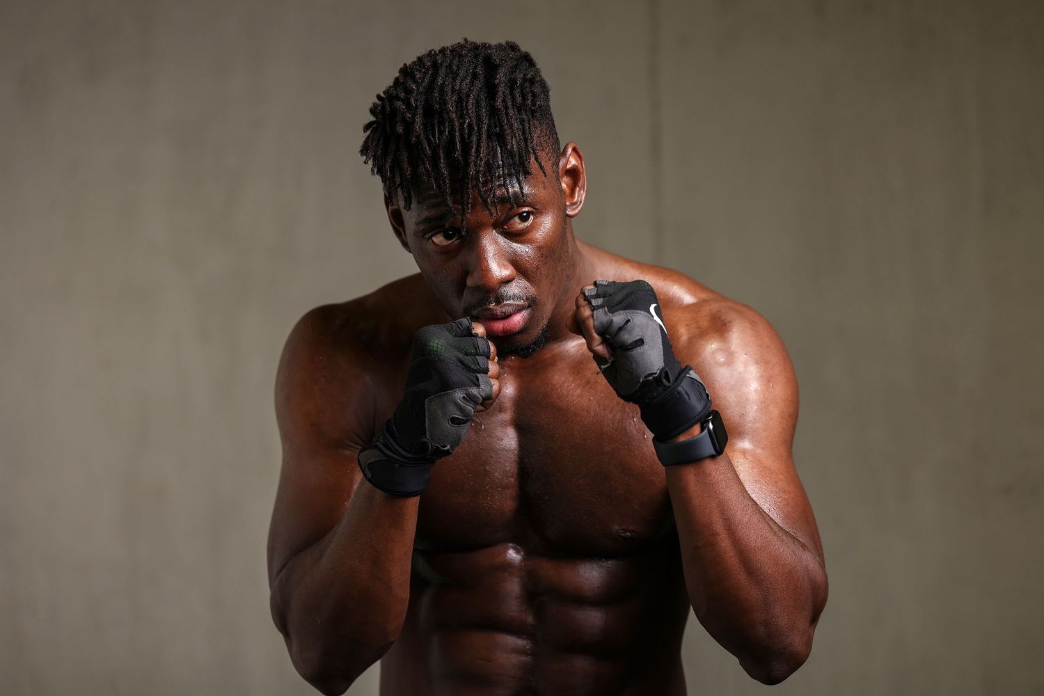 Shirtless muscular Black man in fighting stance wearing boxing gloves against gray background.