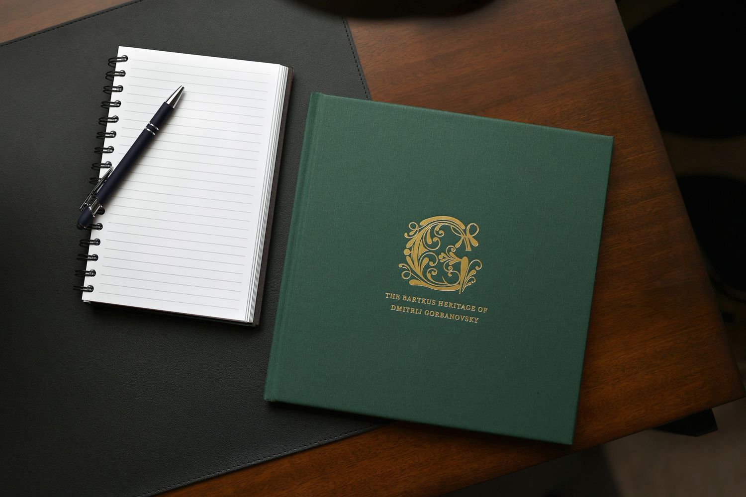 Green hardcover journal beside a spiral notebook and pen on a dark desk.