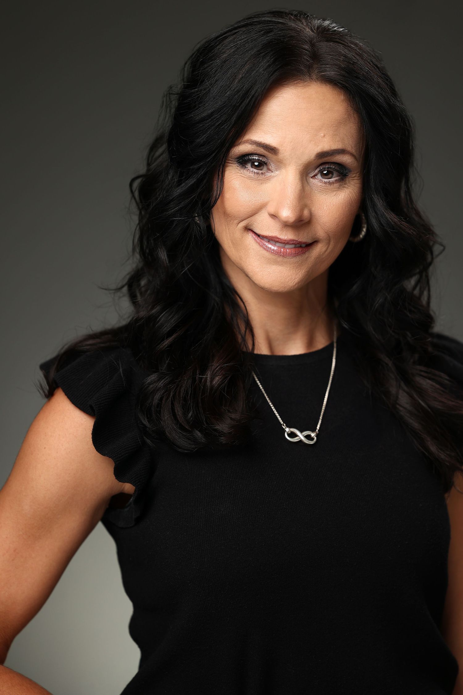 Professional headshot of a smiling woman with dark wavy hair wearing a black dress and infinity necklace.