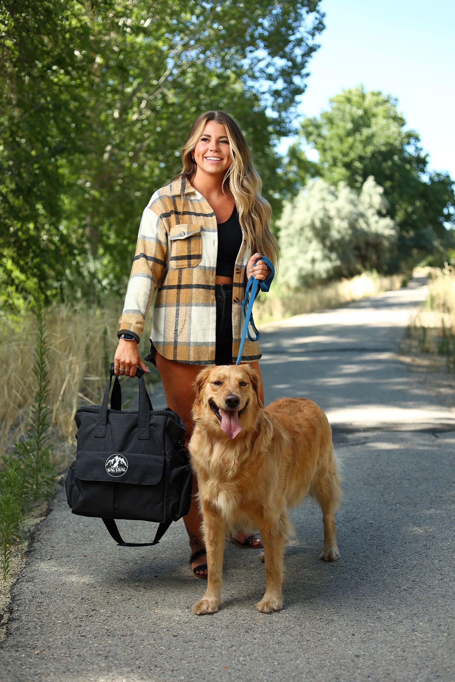 Smiling blonde woman in plaid jacket walking a golden retriever on a tree-lined path while holding a black tote bag.