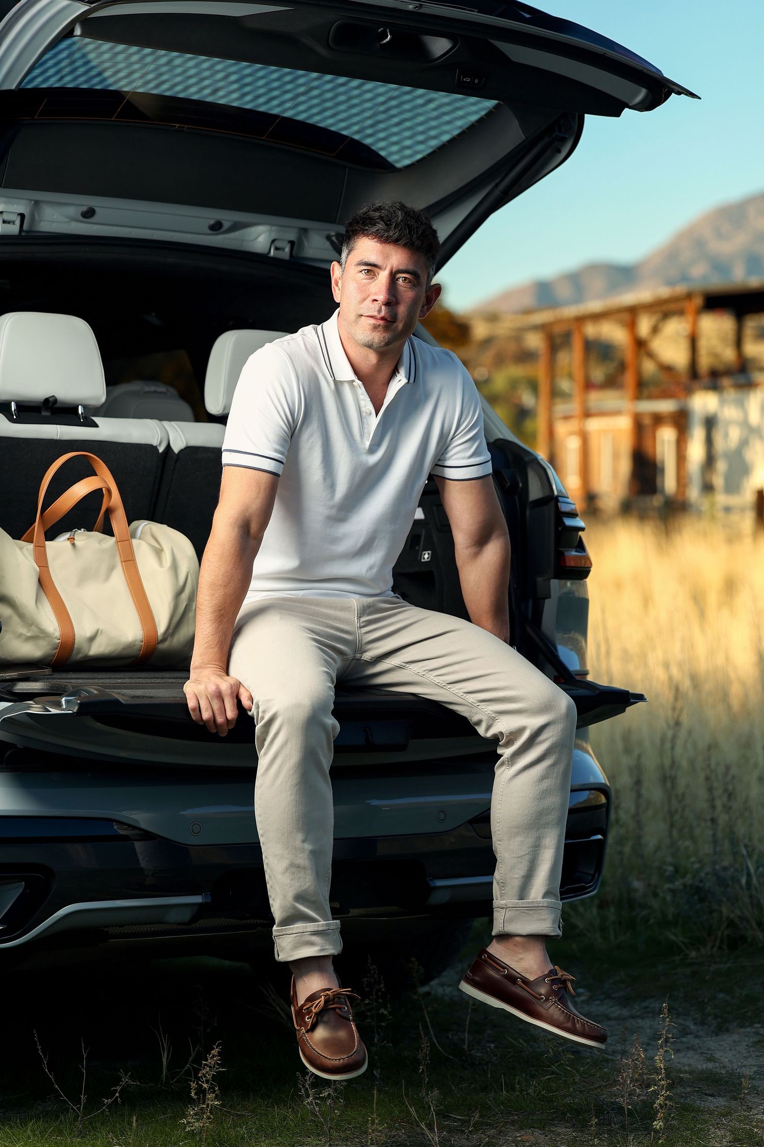 Confident man in white polo shirt and khaki pants sitting on open car trunk in outdoor setting.
