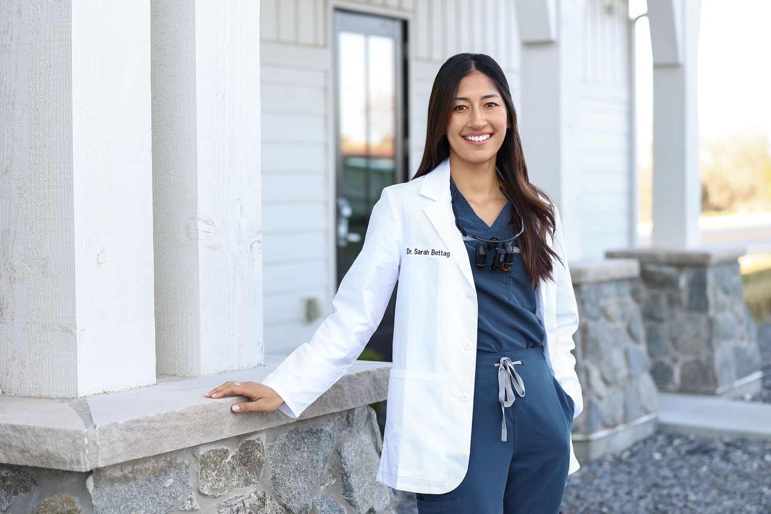 Smiling female doctor in white lab coat and navy scrubs posing outdoors near a building entrance.