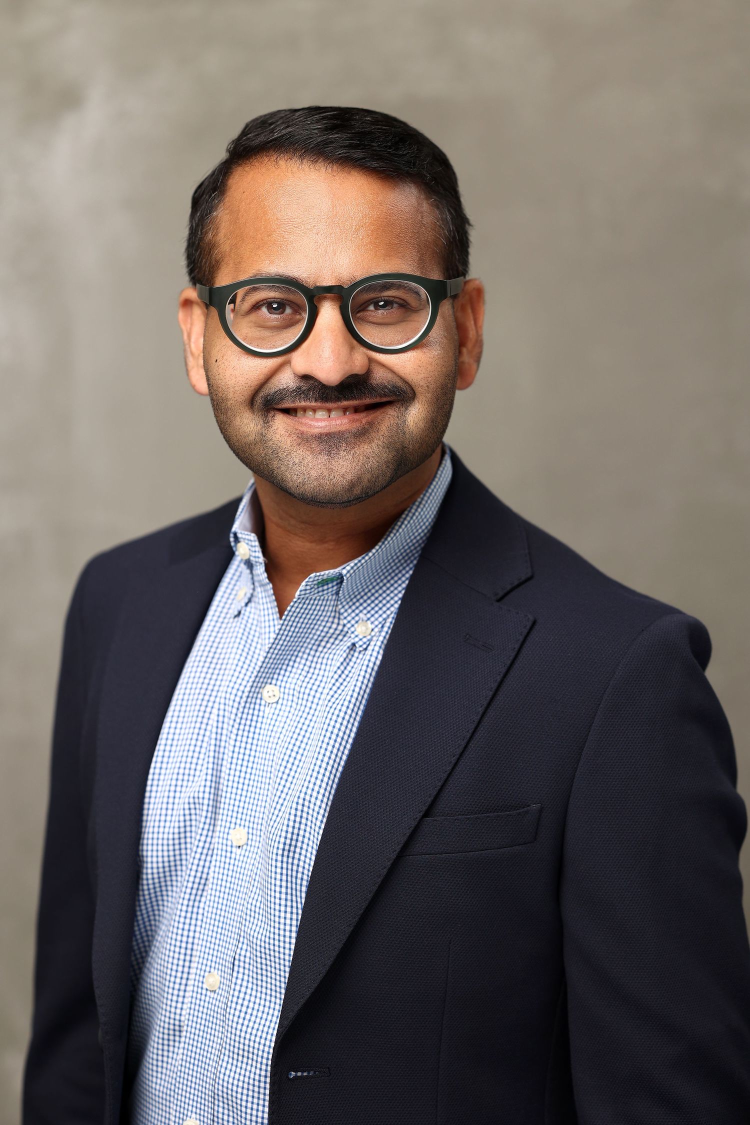 Professional headshot of a smiling man with glasses and a beard wearing a navy blazer and checkered shirt.