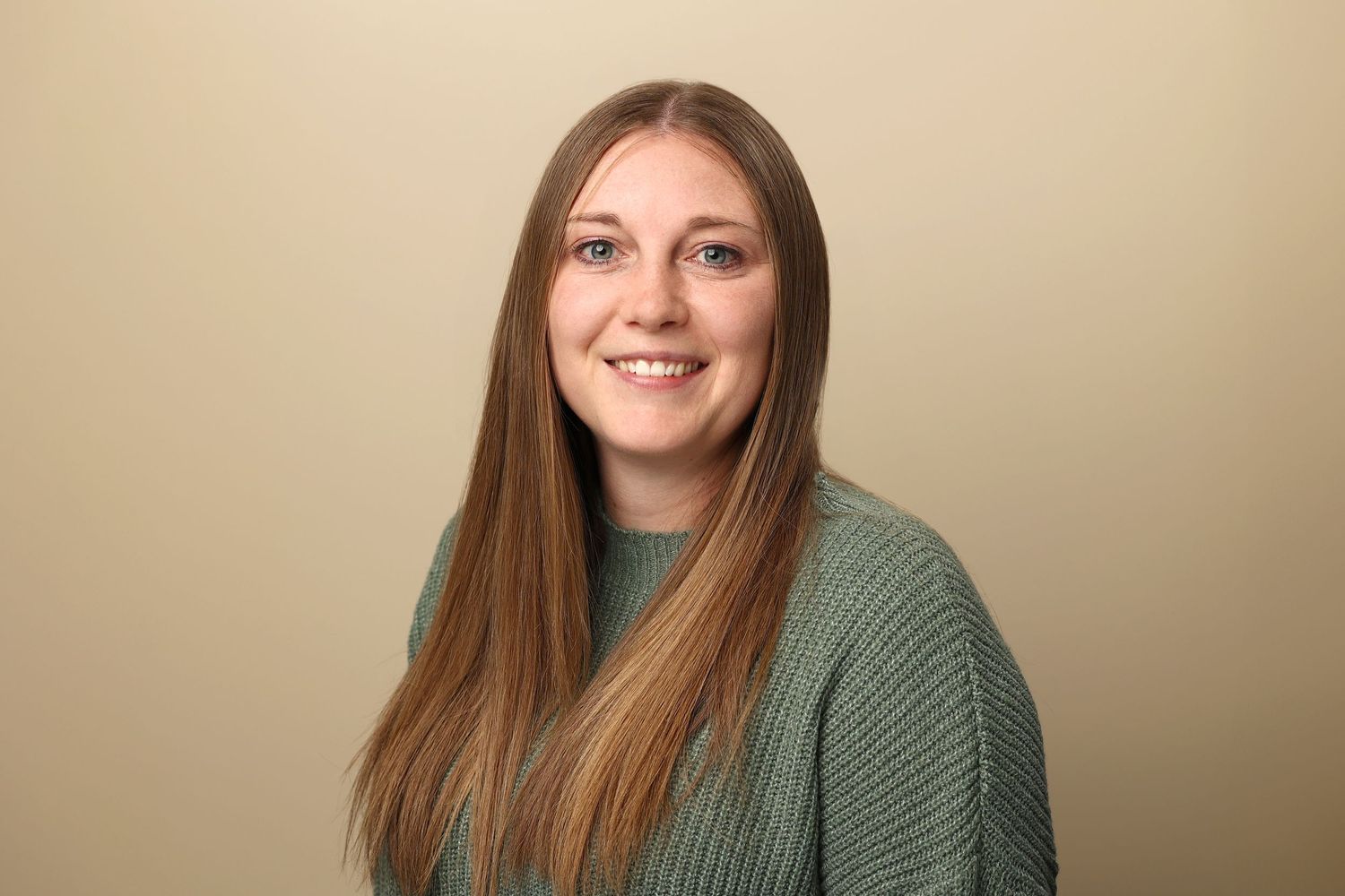 Young woman with long brown hair smiles for professional headshot on beige background.