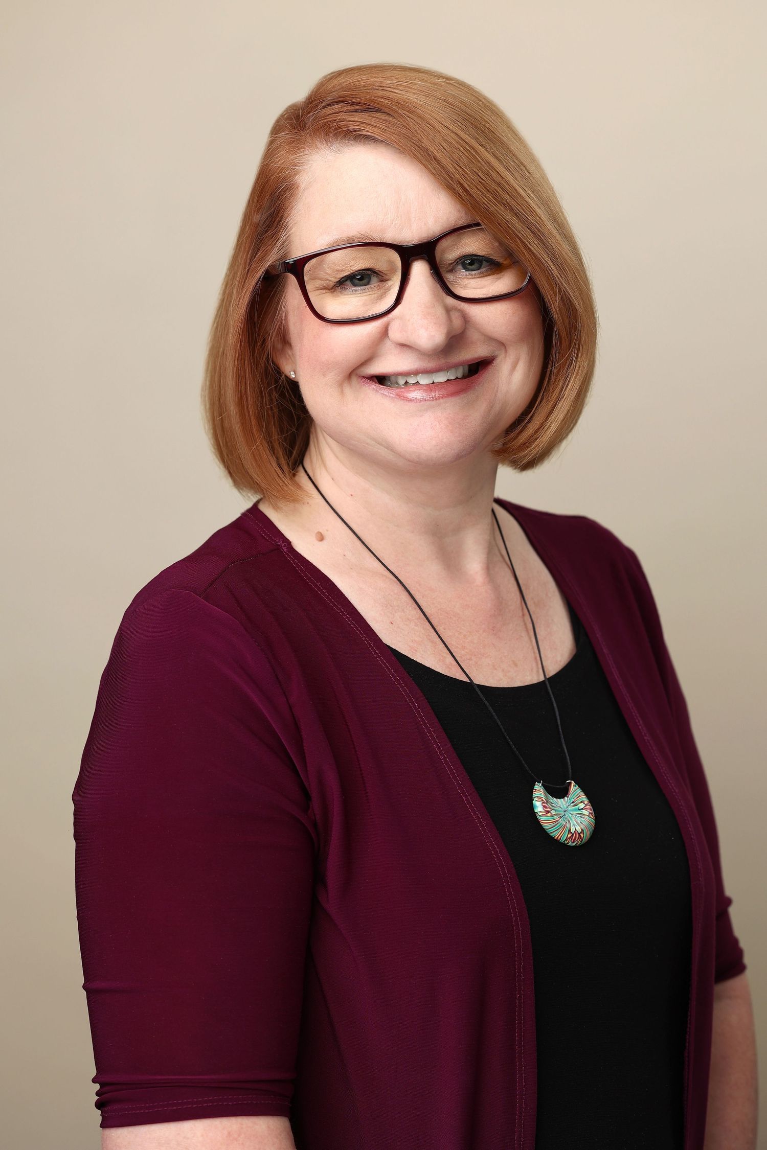 Professional headshot of a smiling woman with red hair and glasses wearing a burgundy cardigan.