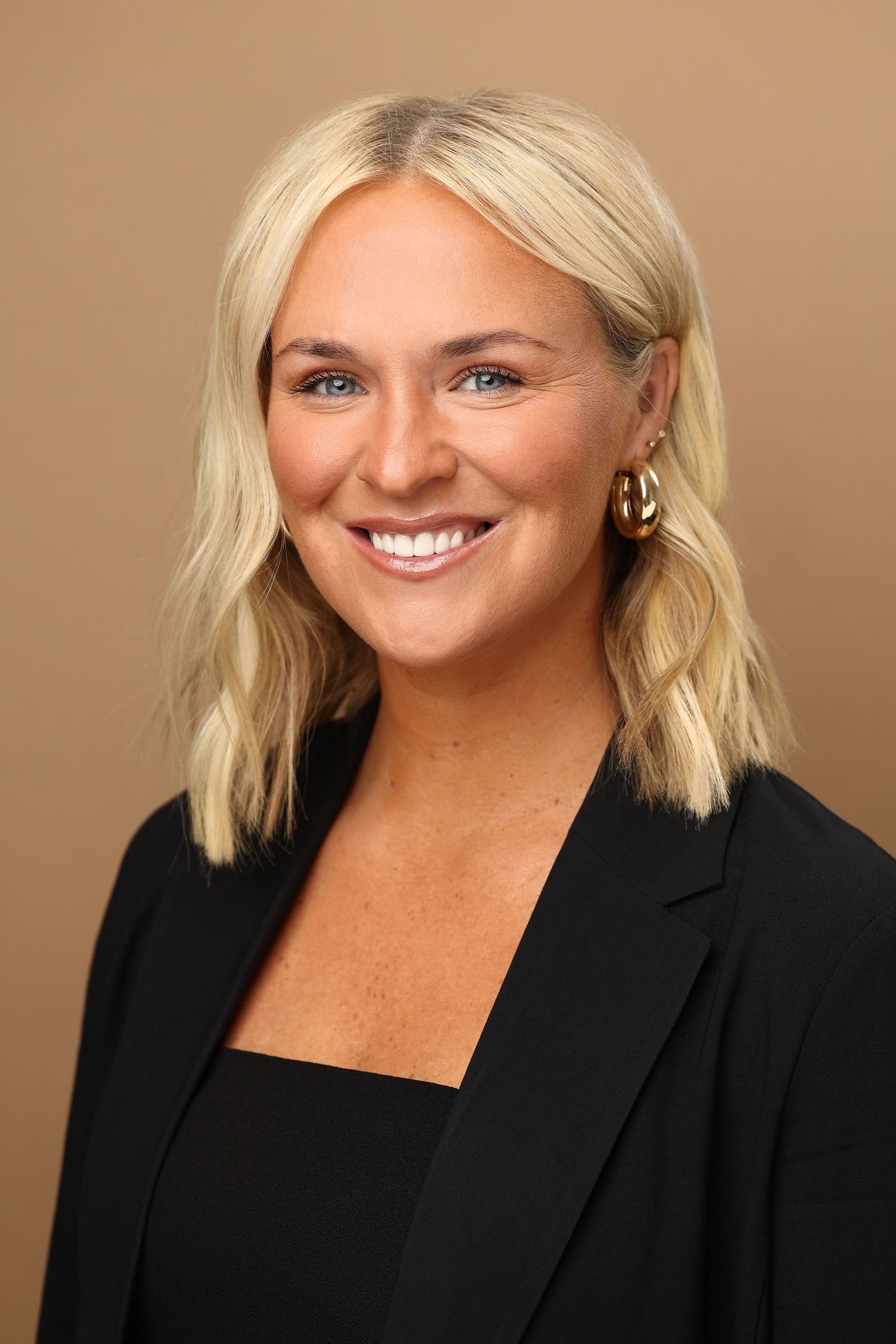 Professional headshot of a smiling blonde woman in a black blazer with gold hoop earrings.