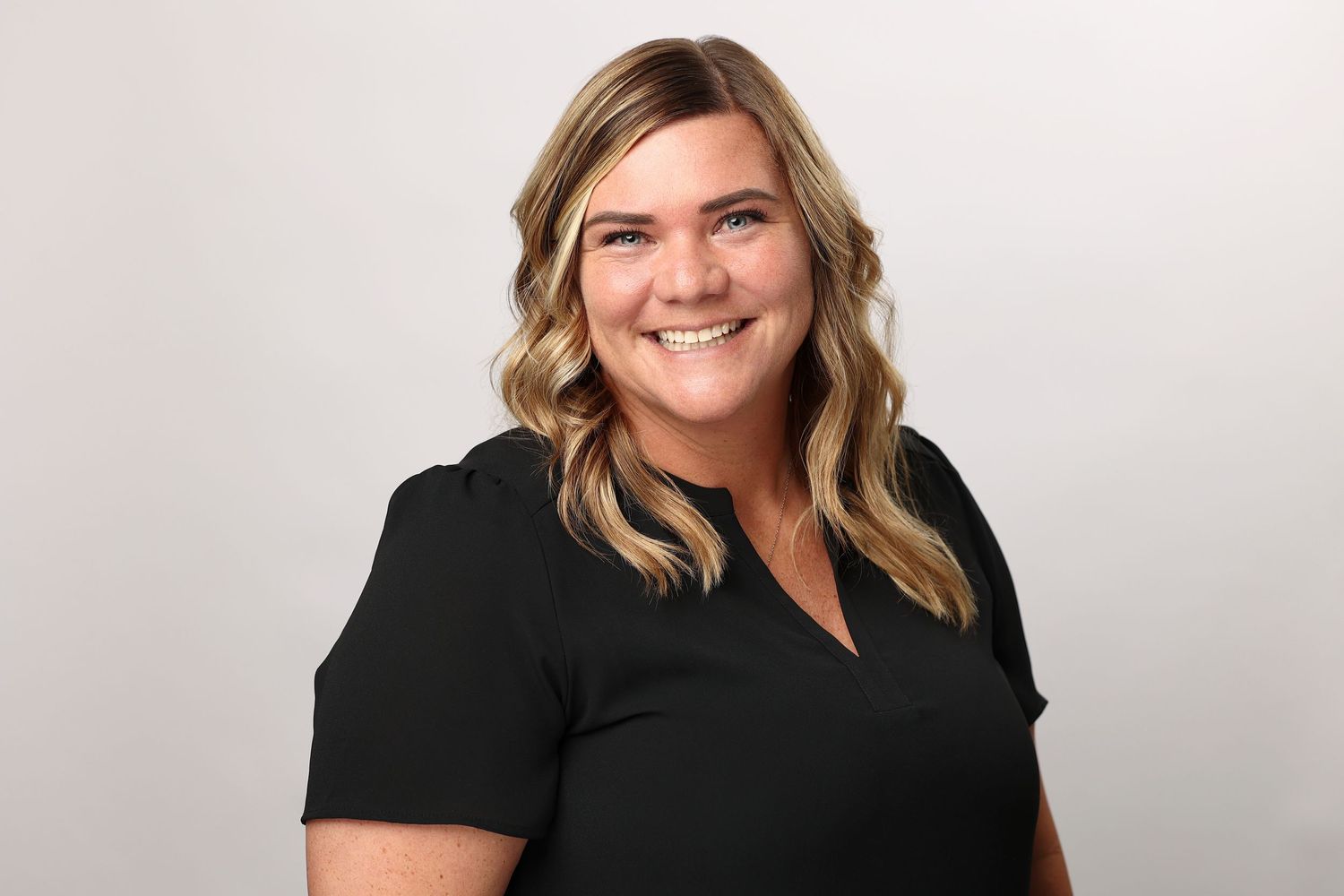 Smiling blonde woman in black top poses for professional headshot on white background.