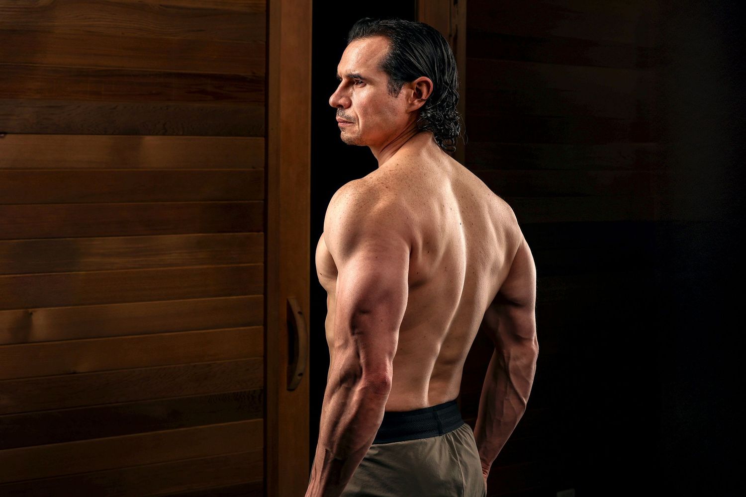 Muscular man showing defined back muscles in dramatic lighting against wooden background.