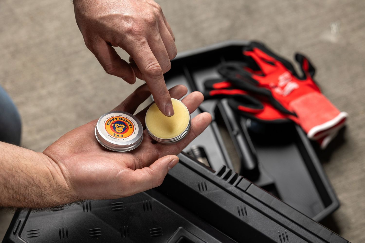 Hands applying Bunky Monkey SAV balm from tin near toolbox and red gloves.