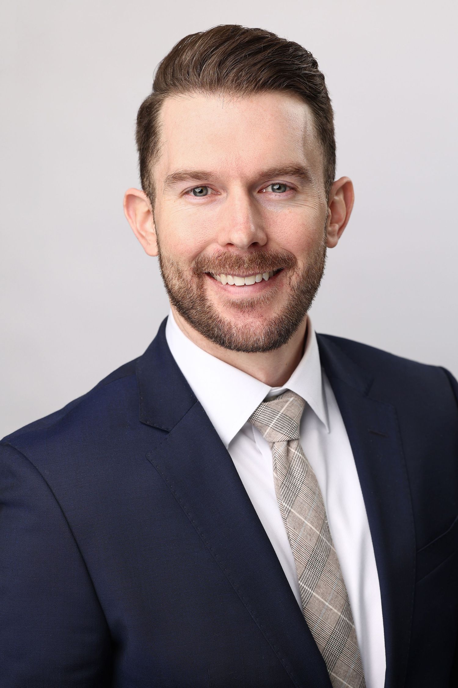 Professional headshot of man in navy suit and tie with beard smiling against light background.