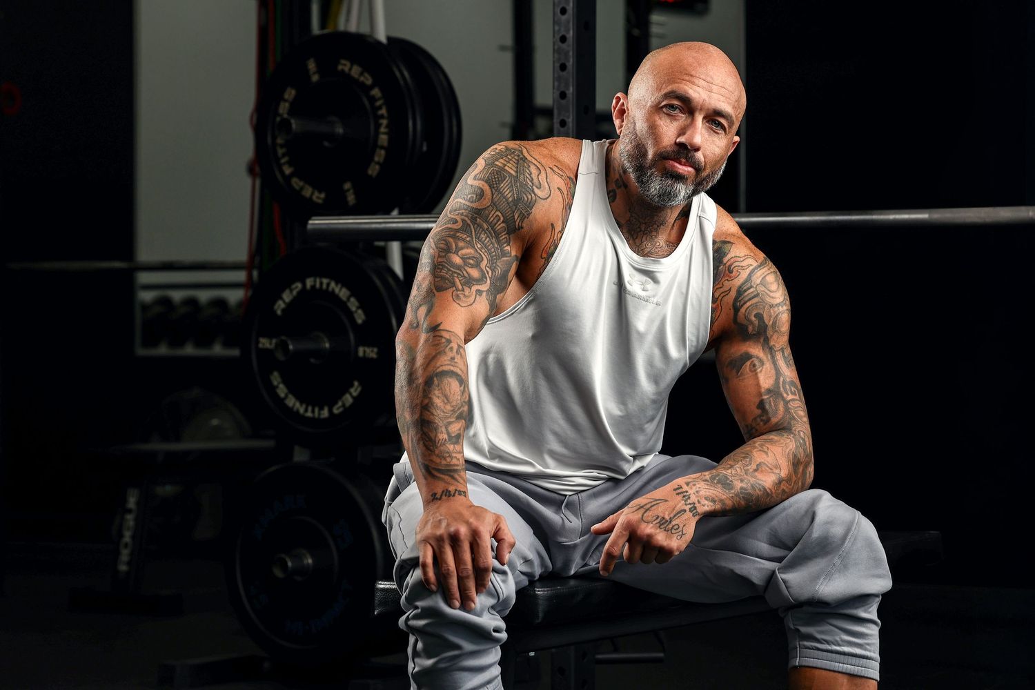 Tattooed bald muscular man sitting in gym wearing white tank top and gray sweatpants.