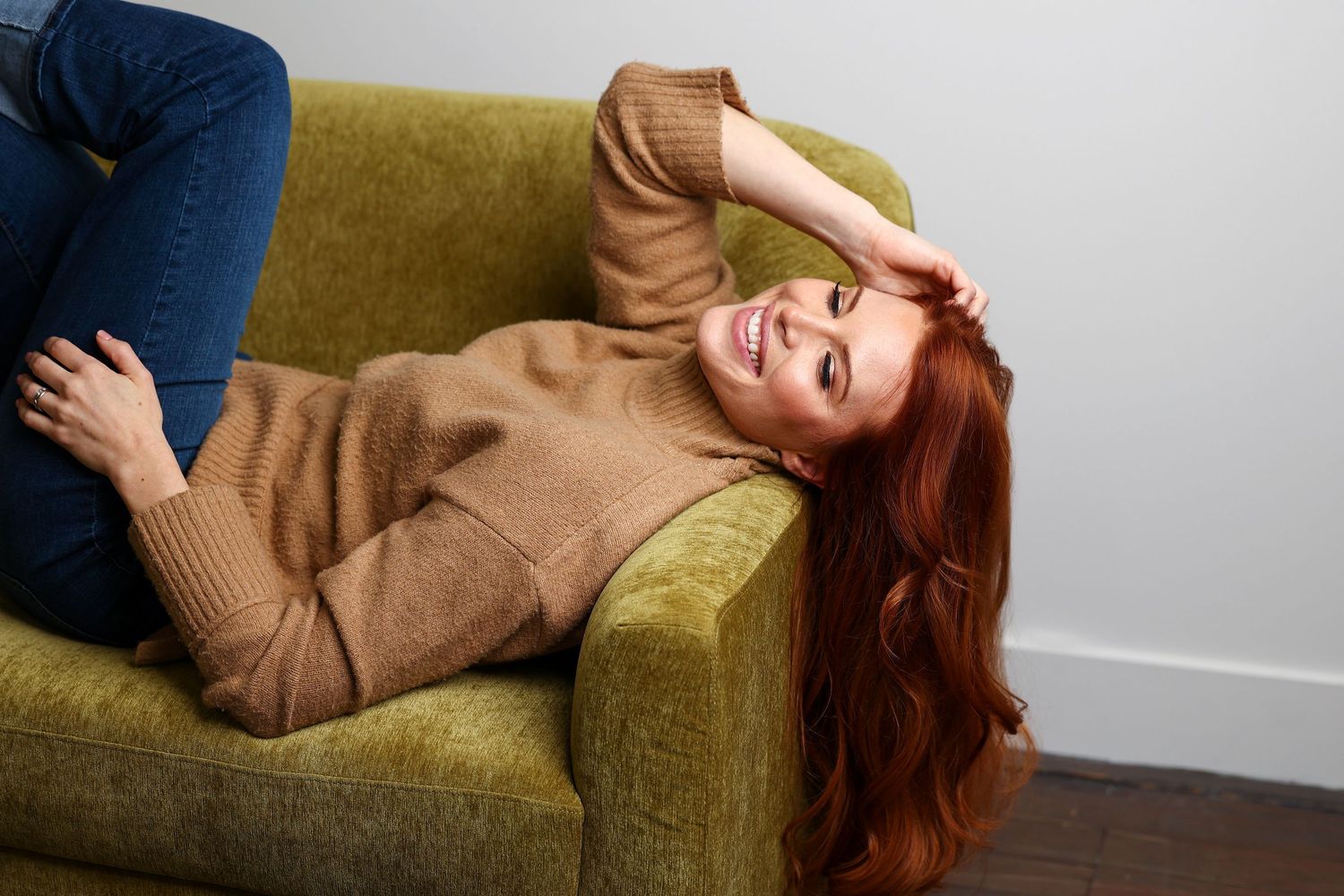 Smiling redhead woman relaxing on a green armchair, wearing a beige sweater and blue jeans.