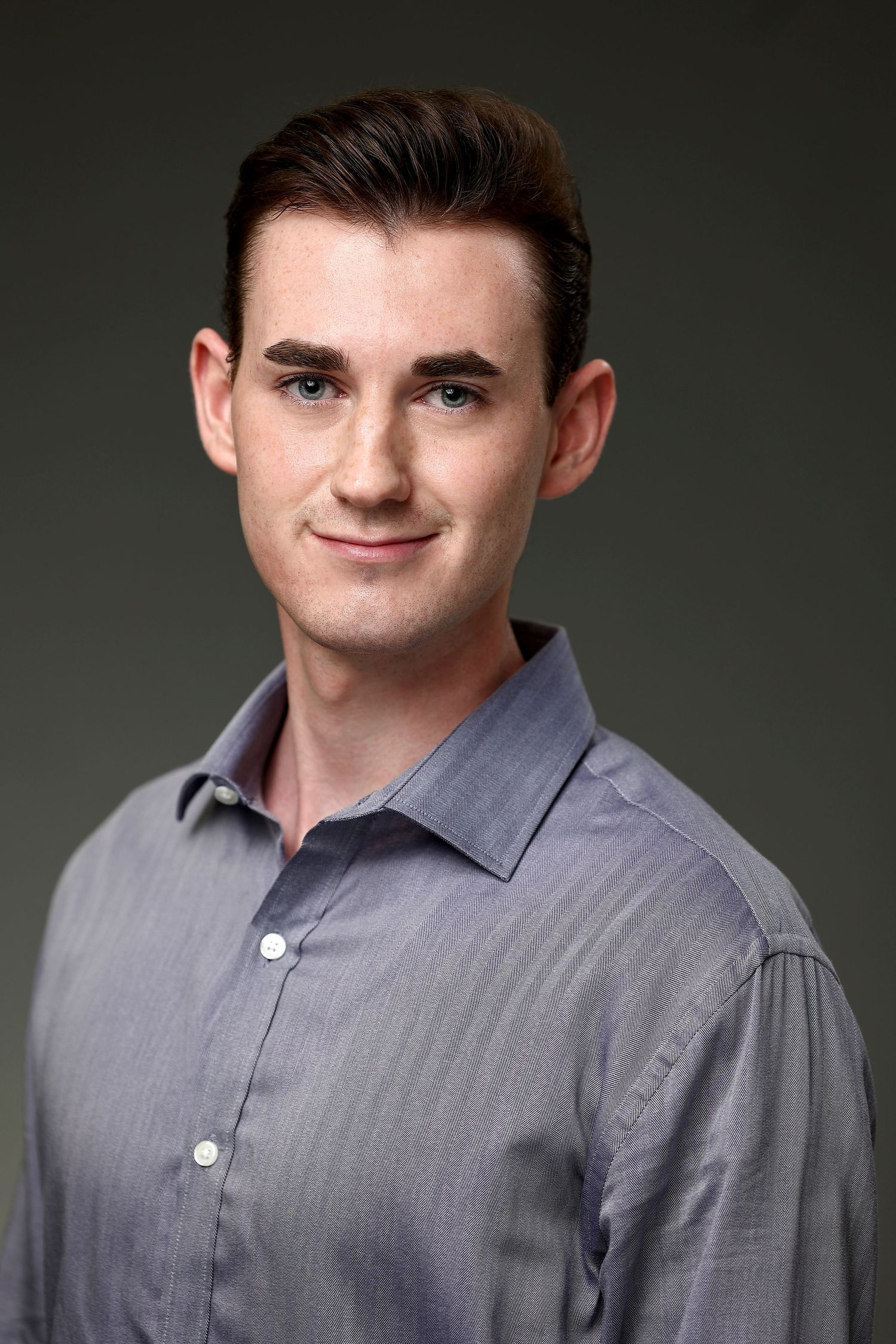 Professional headshot of a young man in a gray button-up shirt against a dark background.