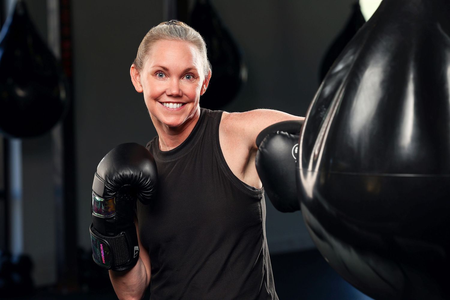 Smiling woman in black sleeveless top boxing with heavy bag in gym wearing gloves.