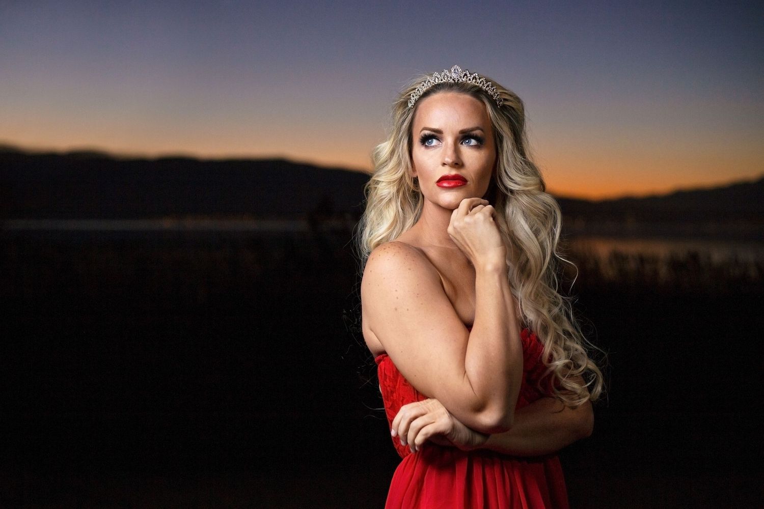 Elegant blonde woman in red gown wearing a tiara at sunset by a lake.