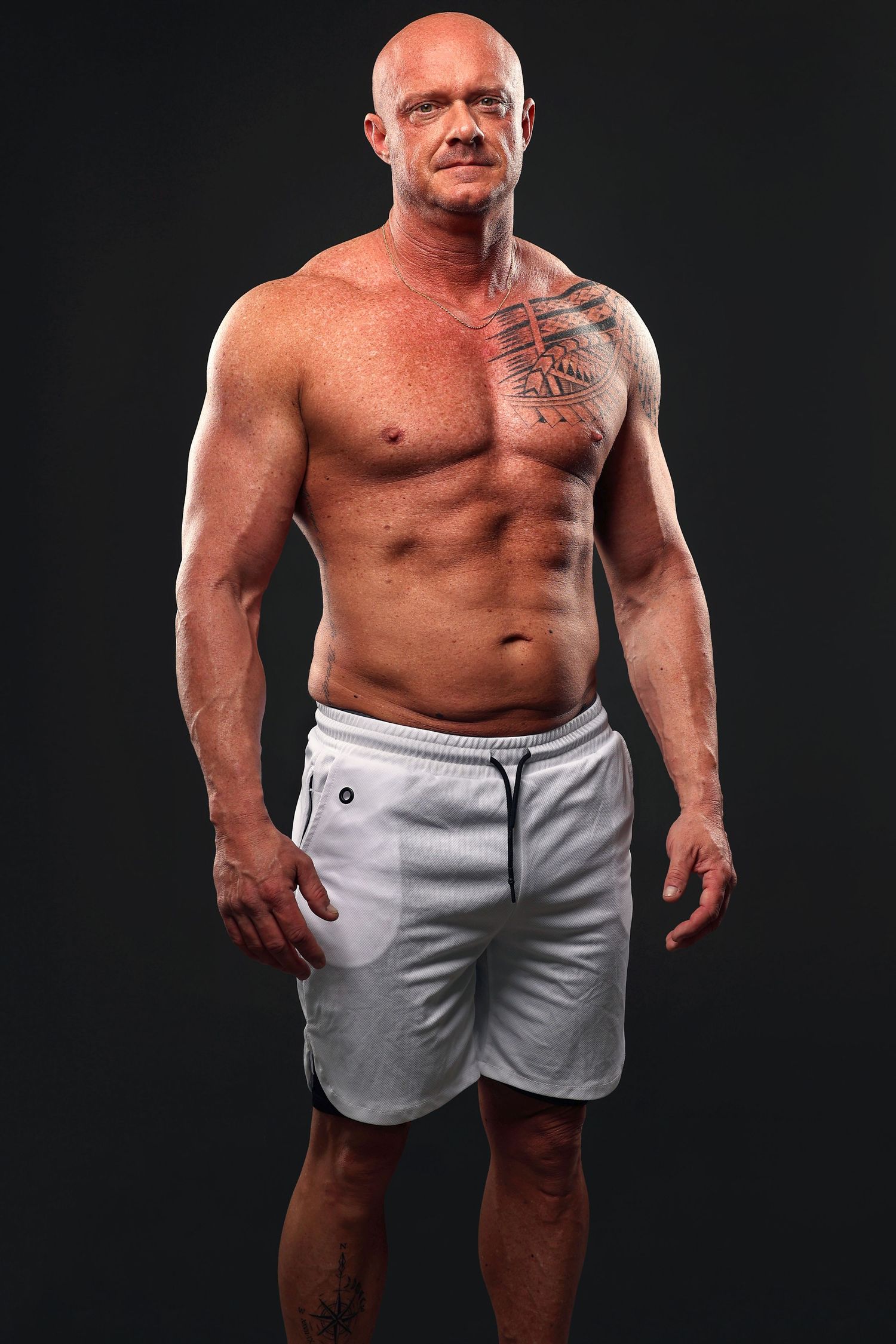 Muscular bald man with chest tattoo posing shirtless in white shorts against dark background.