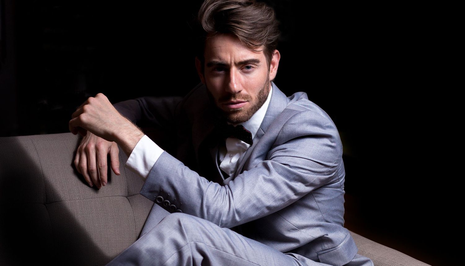 Stylish man in gray suit sitting in dramatic low-key lighting with serious expression.