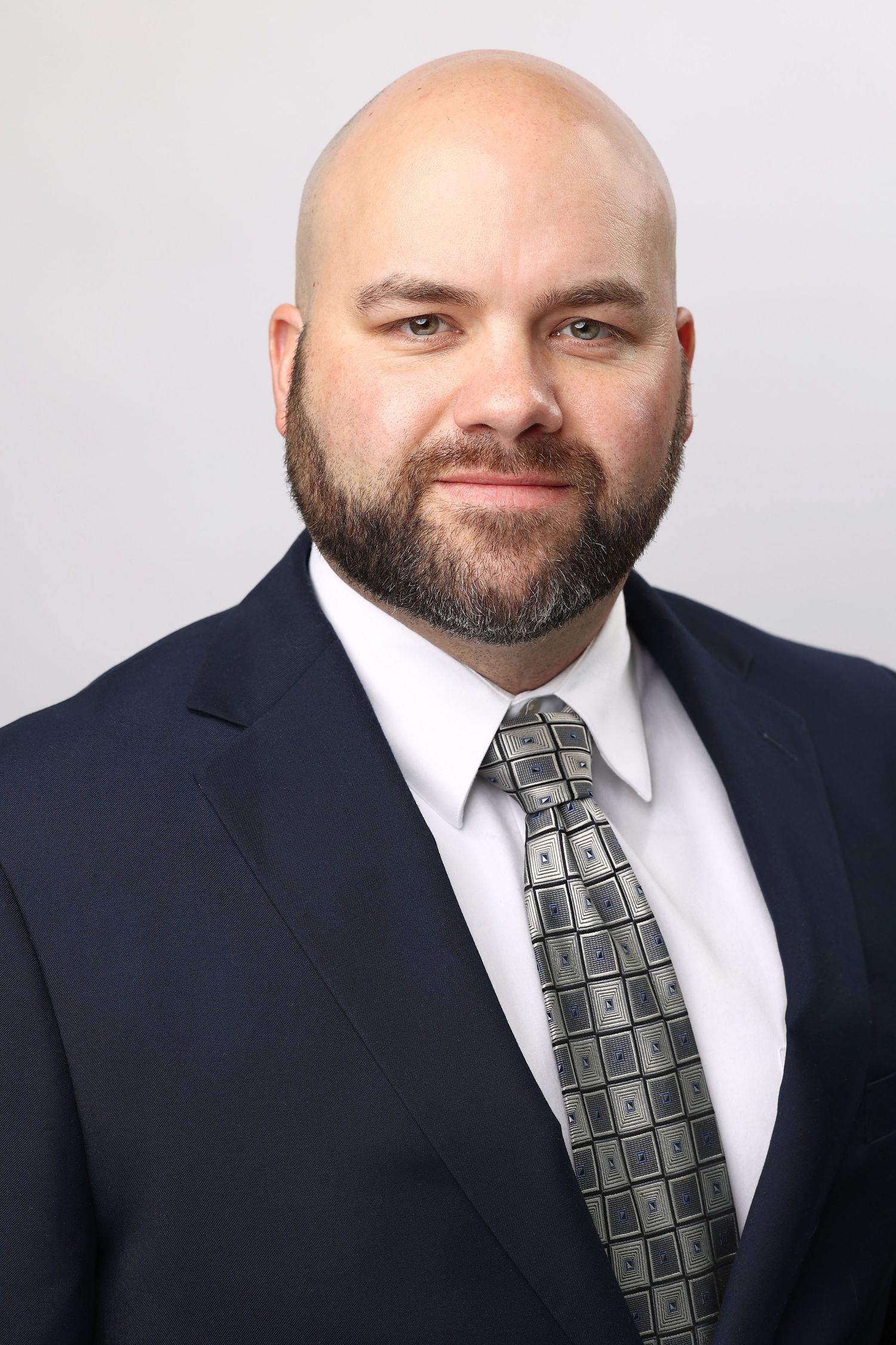 Professional headshot of a bald man with a beard wearing a navy suit and patterned tie.