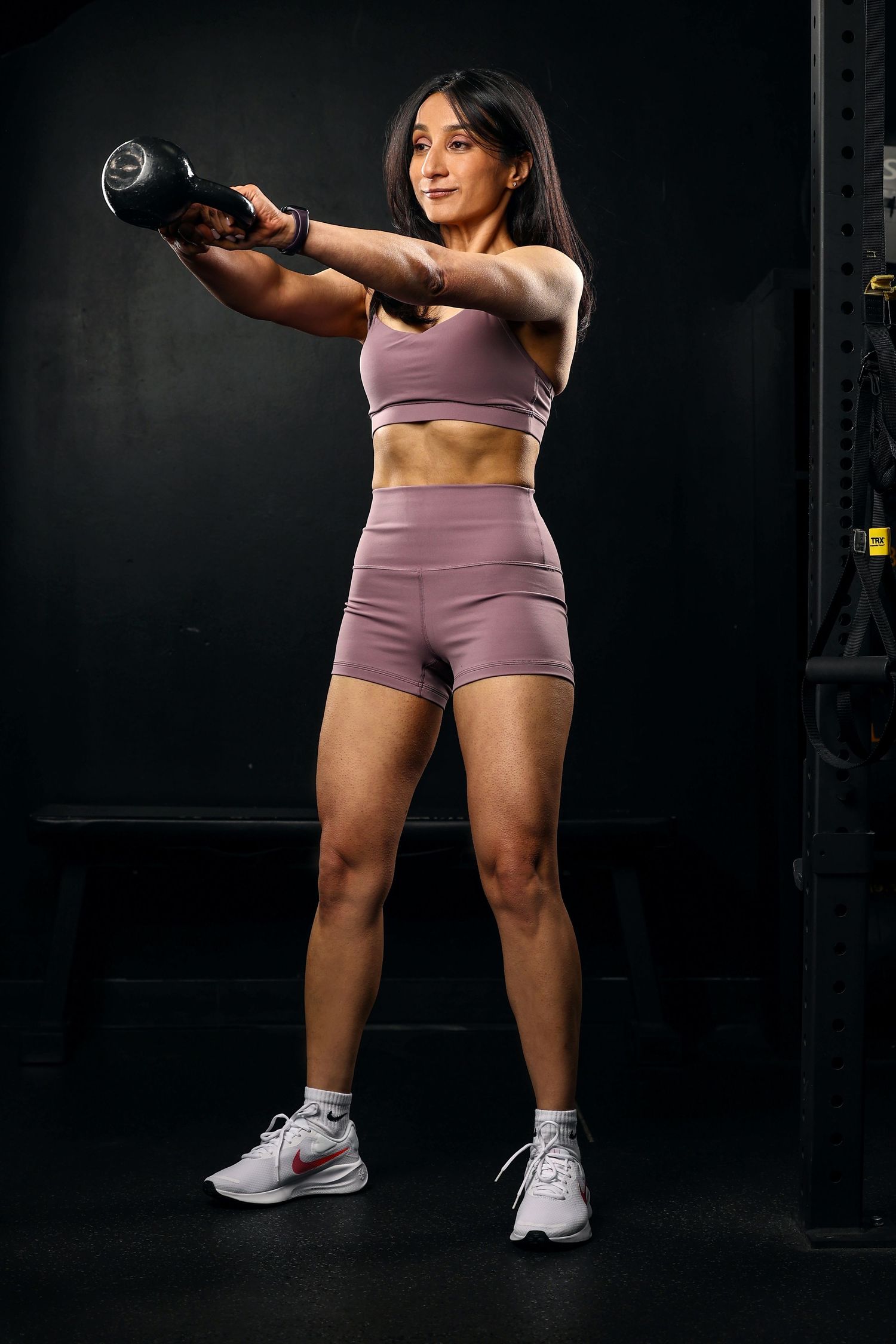 Athletic woman in purple workout set performing dumbbell front raise exercise in dark gym.