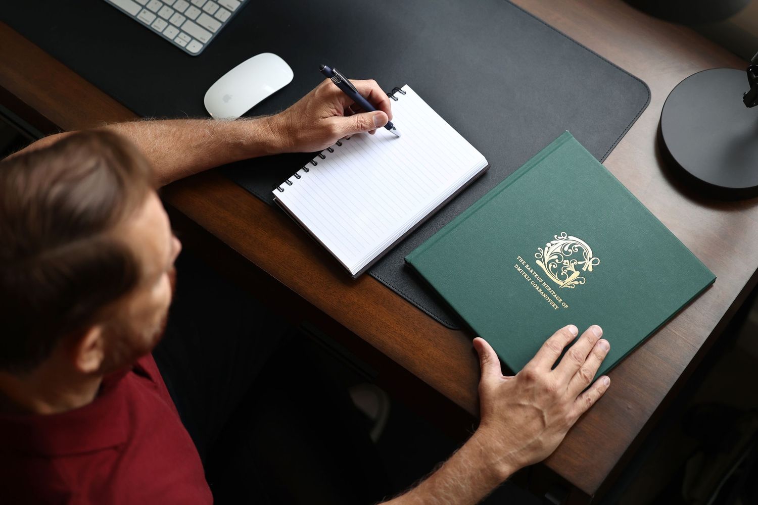 Man writing in a spiral notebook beside a green official document folder at a desk.