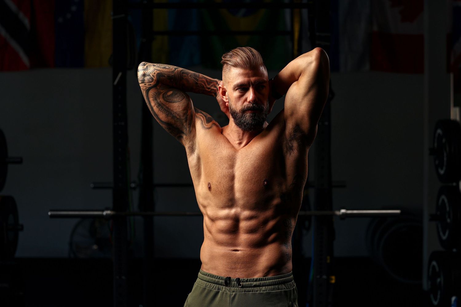 Bearded tattooed shirtless man flexing muscles in dark gym with barbells in background.