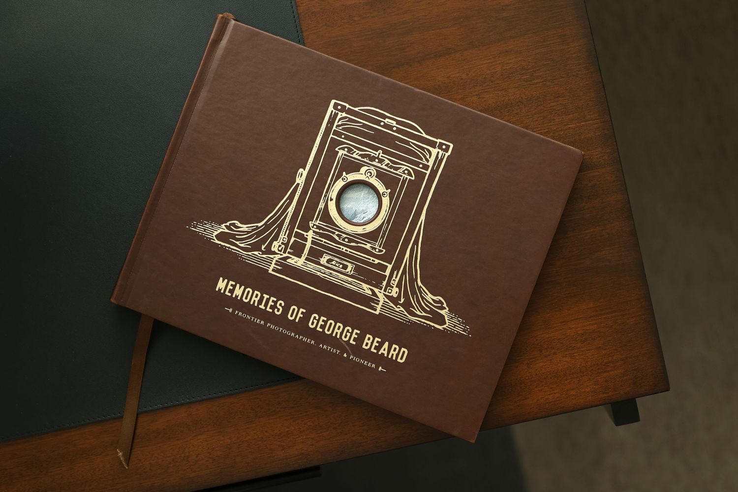 Brown hardcover Memories of George Beard book with gold vintage camera illustration on desk.