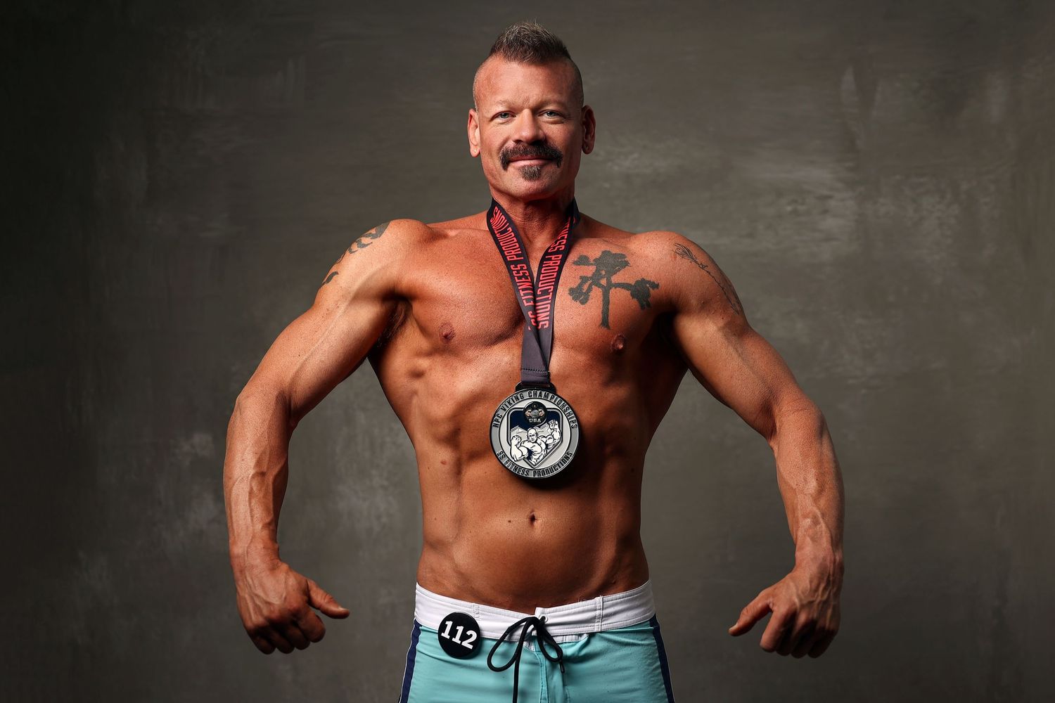 Muscular bodybuilder flexing shirtless with competition medal and chest tattoo on gray background.