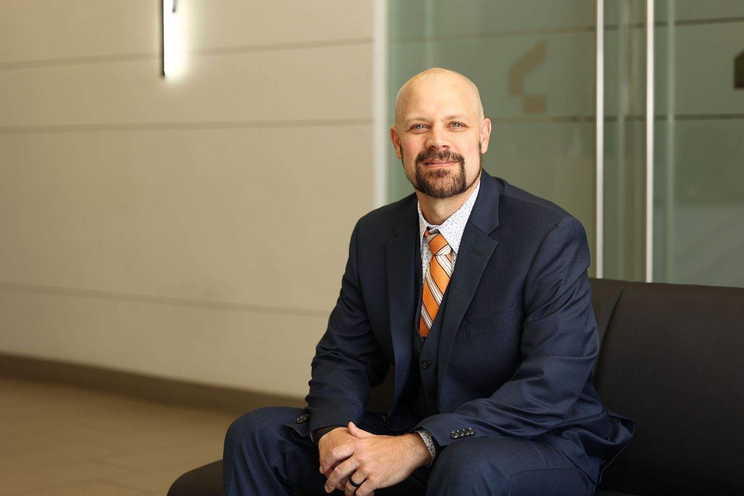 Professional man in blue suit and orange tie seated in modern office lobby.