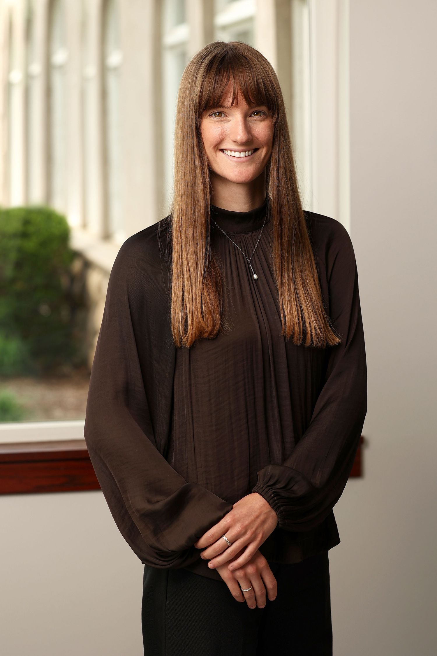 Professional headshot of a tall woman with long brown hair wearing a black blouse standing near a window.