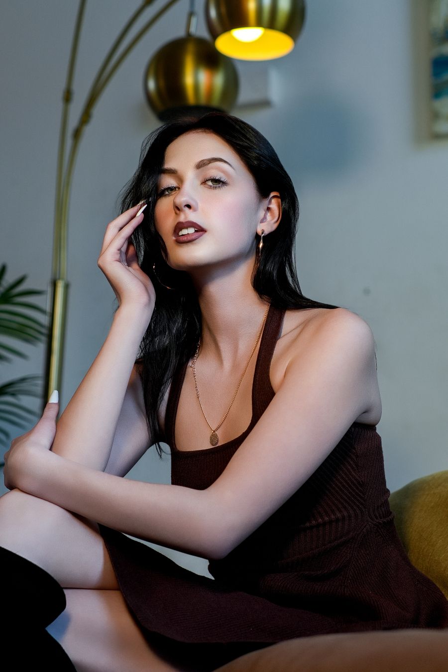 Brunette woman in brown dress poses elegantly indoors under warm gold pendant lamp.