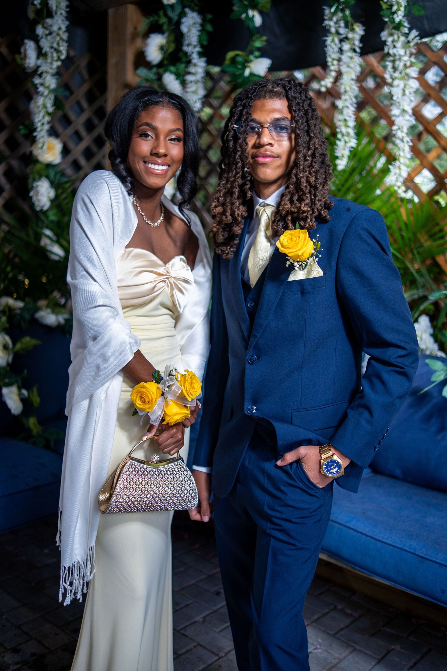 A young couple dressed in prom attire, wearing yellow roses, posing together in front of floral decorations.