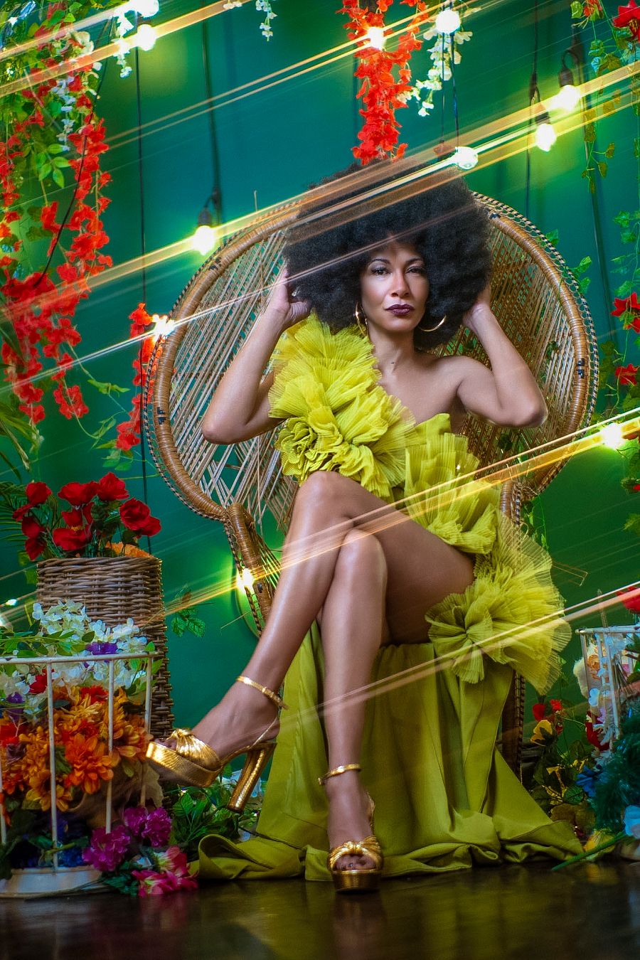 Glamorous woman in yellow ruffle dress poses in peacock chair with vibrant floral decor and warm string lights.
