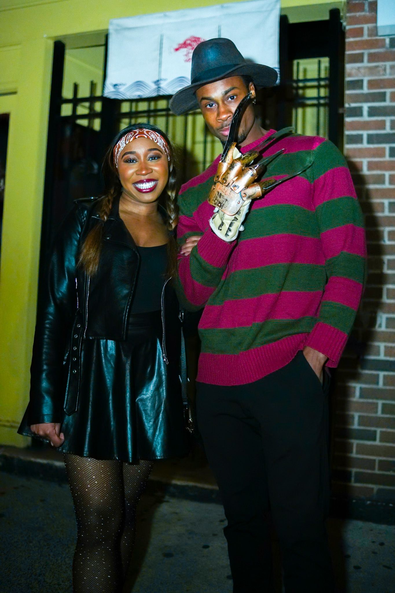 Couple in Halloween costumes, woman in leather skirt and man dressed as Freddy Krueger, pose outdoors.